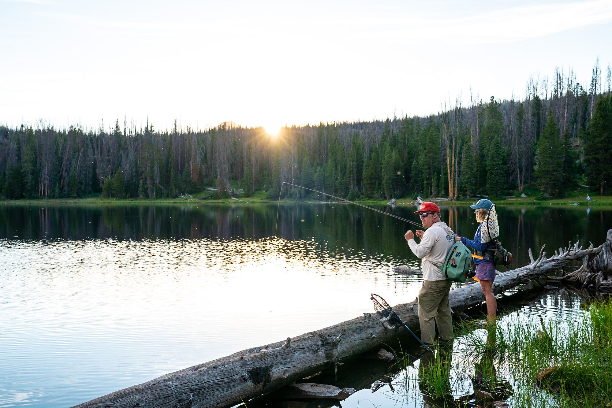 Two people fishing from a log over a lake with a sunset in the background in the Uinta National Forest